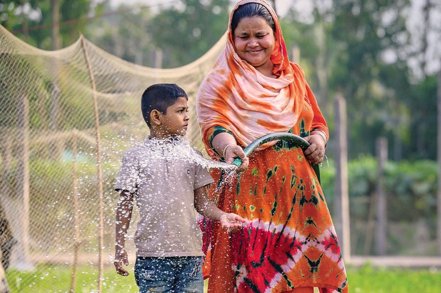 A woman wearing an orange dress waters her vegetables with a young boy beside her. (Bangladesh, 2025)