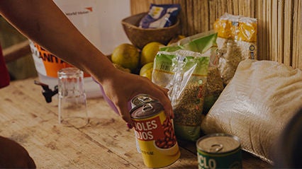 Hand reaches for can of beans amongst other food basket items.