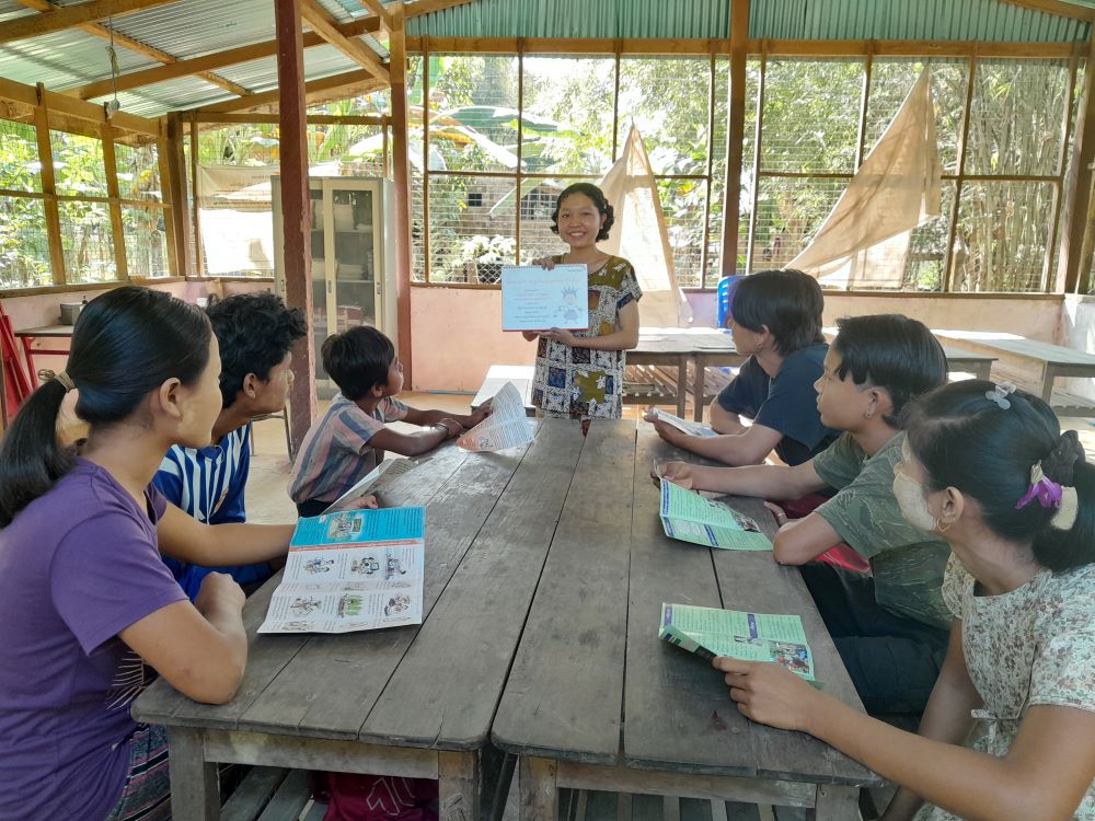 A smiling young girl stands at the head of a table as she helps a group of children with their reading. 
