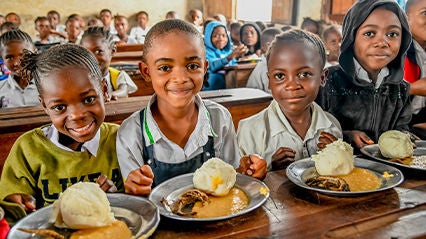Three children smile in front of their plates of food in a classroom.