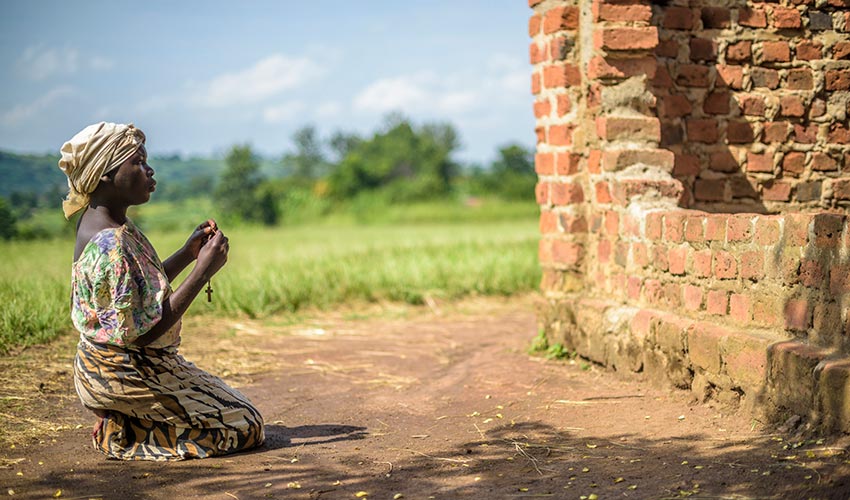 A woman kneels in the dirt outside a brick building with a cross in her hands.