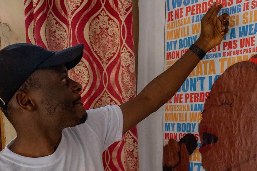A man in a baseball cap, points at a poster on the wall.