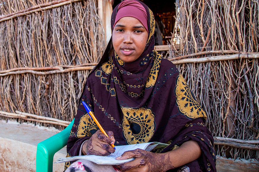 Habiba wearing a hijab sitting in front of her house writing on a notebook.