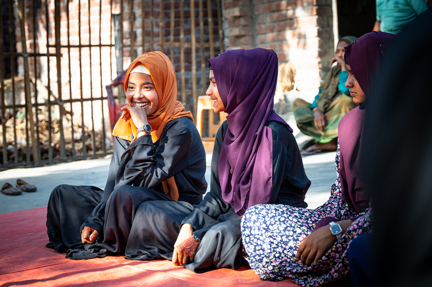 Sultana laughs, sitting on mats with a few other girls. She is wearing an orange hijab.