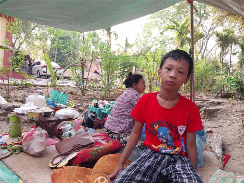A school-aged boy crouches under a tarpaulin shelter amidst tropical vegetation. 