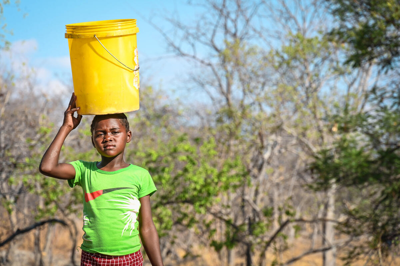 A young girl is pictured carrying a yellow bucket of water over her head.