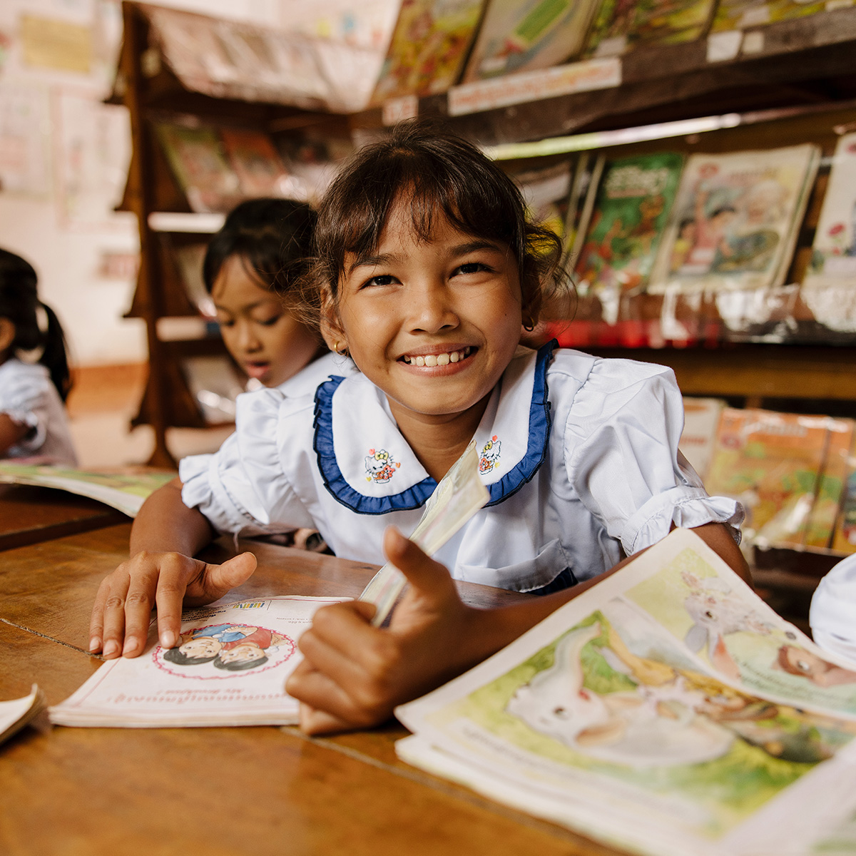 A young girl in a classroom smiles for the camera with a book in front of her.