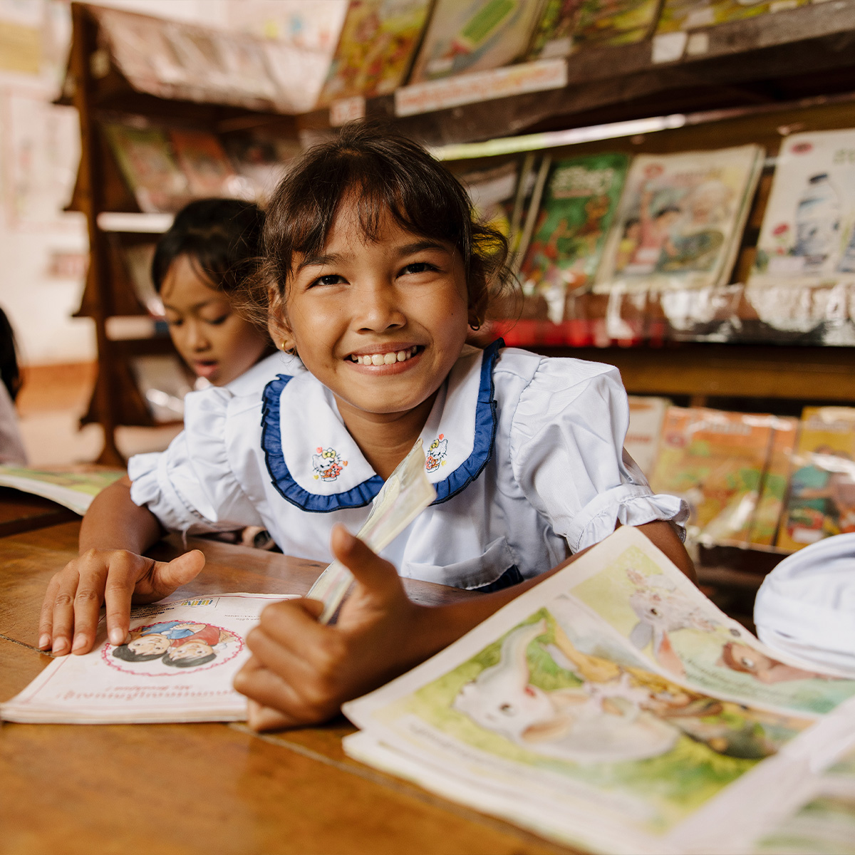 A young girl in a classroom smiles for the camera with a book in front of her.