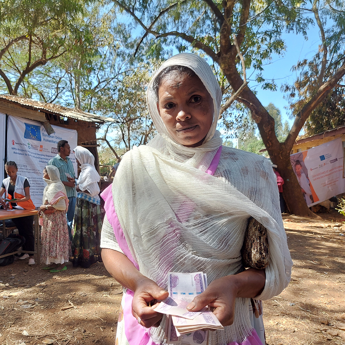 A woman looks at the camera, holding cash. Behind her, people wait in line to receive cash and vouchers.