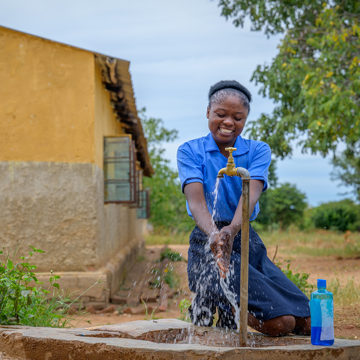 A smiling girl in a blue shirt kneels by an outdoor faucet, washing her hands.