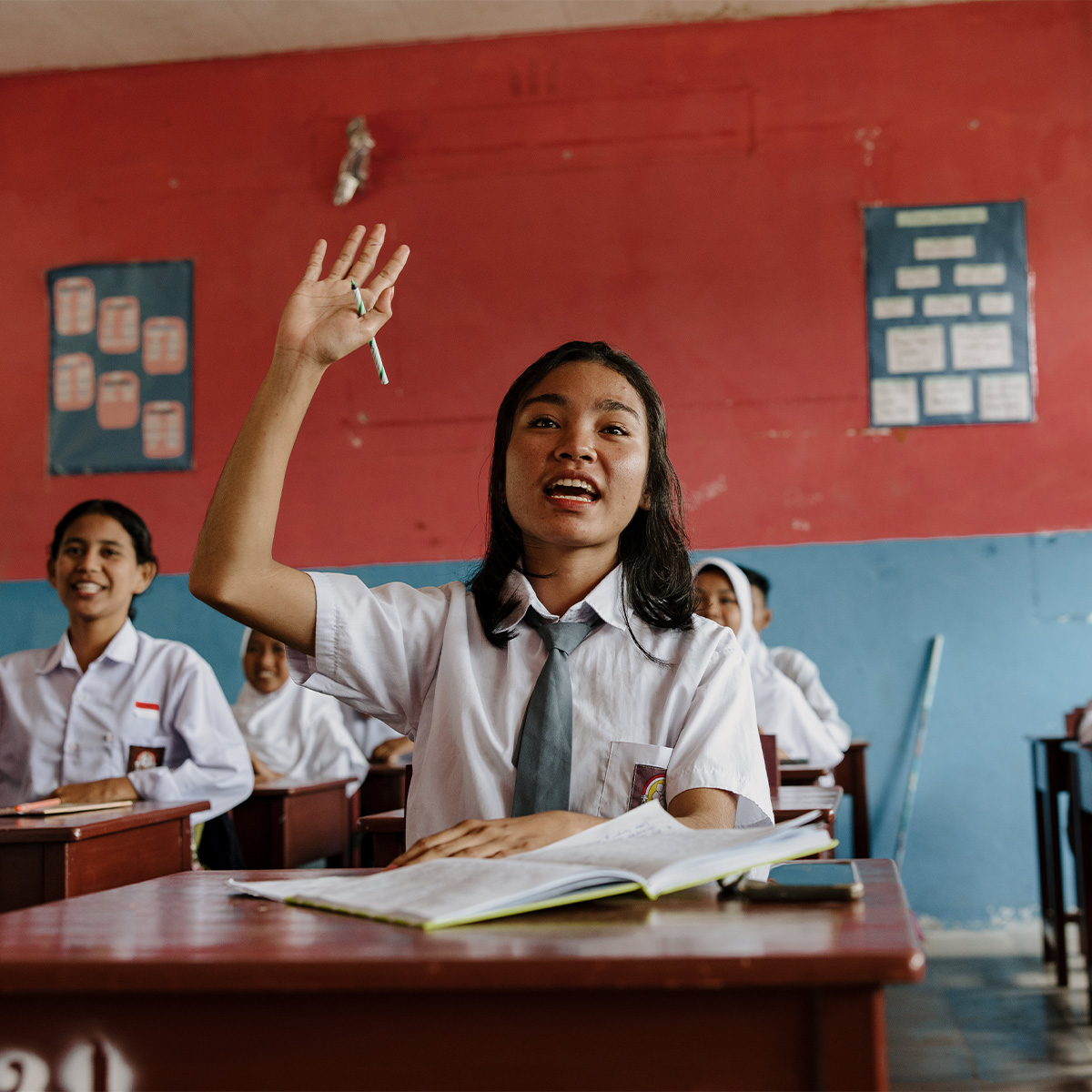 A classroom of girls in uniforms sit at wooden desks, with one girl raising her hand towards her teacher.