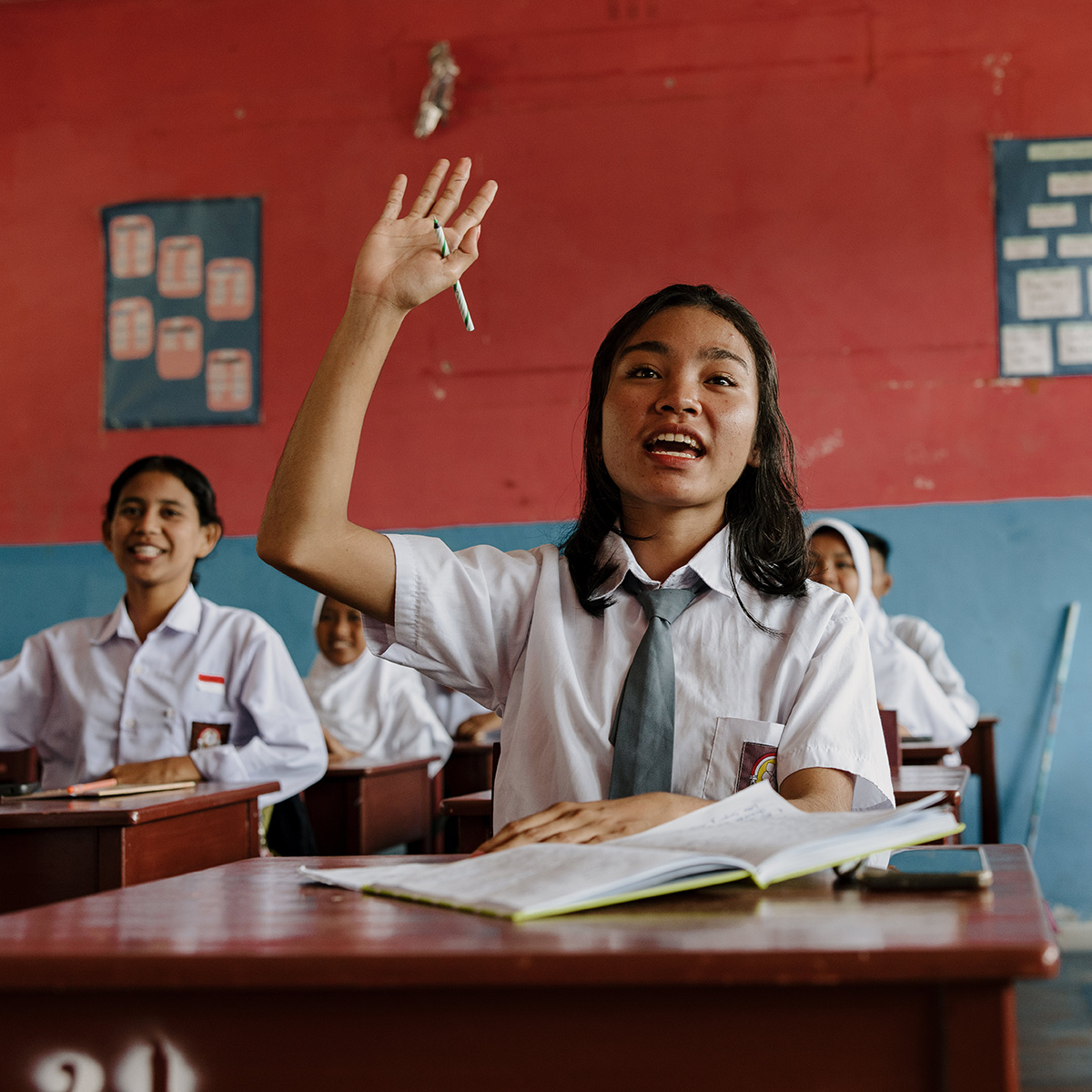 A classroom of girls in uniforms sit at wooden desks, with one girl raising her hand towards her teacher.