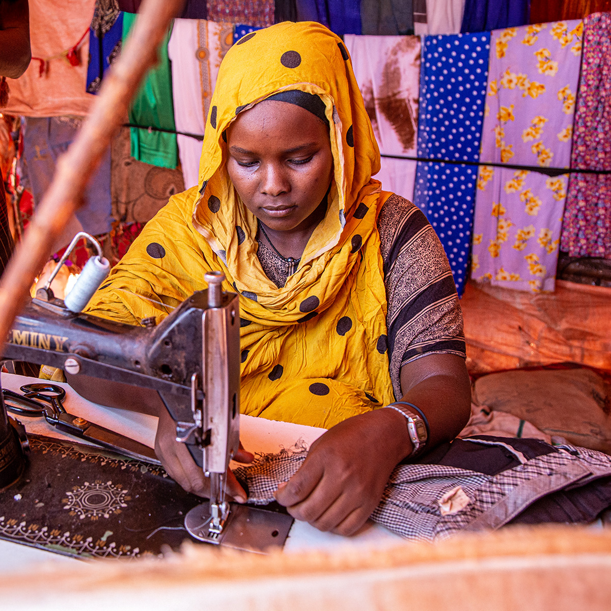 A woman wearing a yellow headscarf works at a sewing machine, surrounded by colorful fabrics.