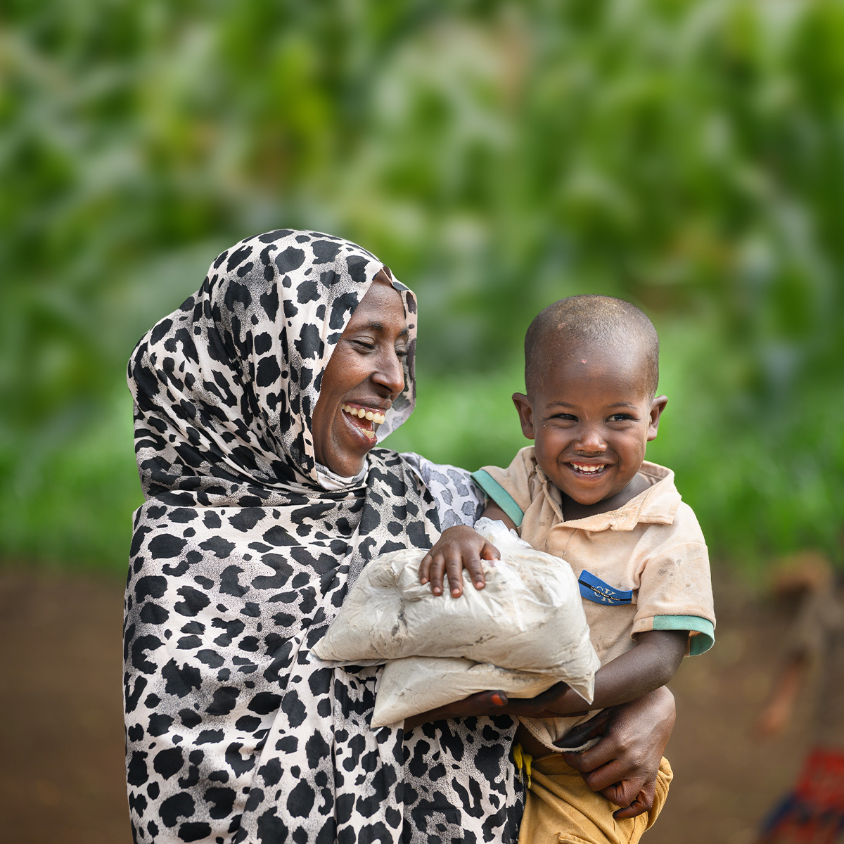 A woman is wearing patterned clothing holding a small child and a package of food supplies outdoors.