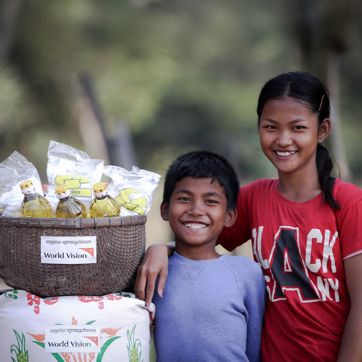 A sister places her hand over her brother's shoulders as they both smile while standing next to essential food items provided by World Vision.