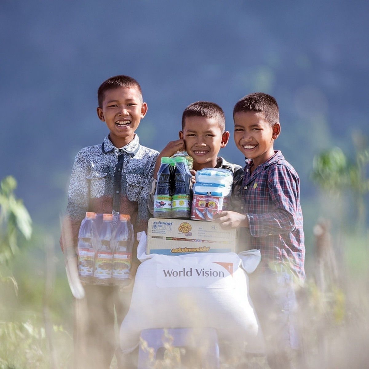 Three boys smile next to each other as they stand behind a pile of food items.