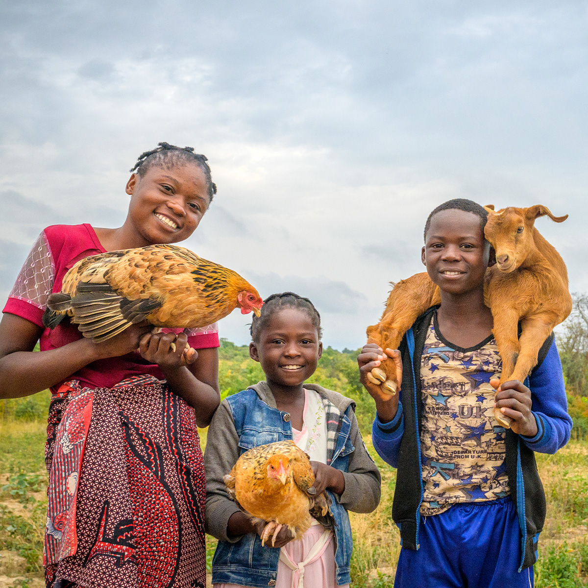 Two girls hold chickens in their arms next to a boy holding a fluffy goat around his shoulders.