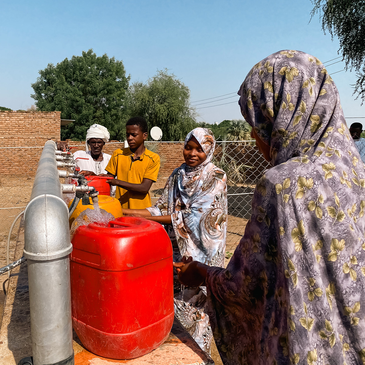 Two women and two men fill up red jugs with water at a water pumping station.