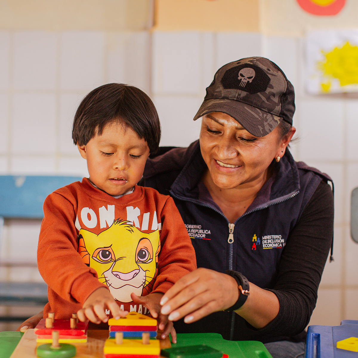 A woman assists a young boy with stacking colourful blocks on pegs at a table.