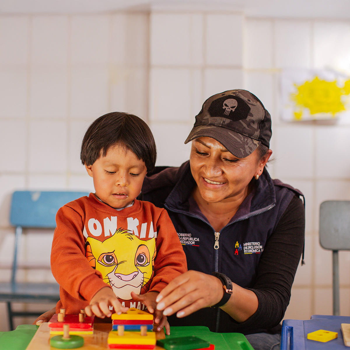 A woman assists a young boy with stacking colourful blocks on pegs at a table.