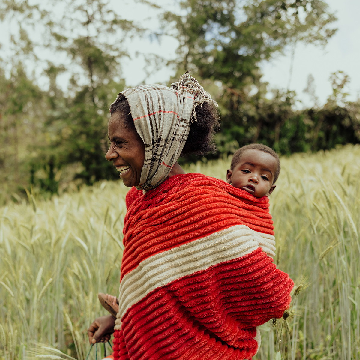 A smiling woman wears her baby on her back, with the baby peeking over her shoulder.