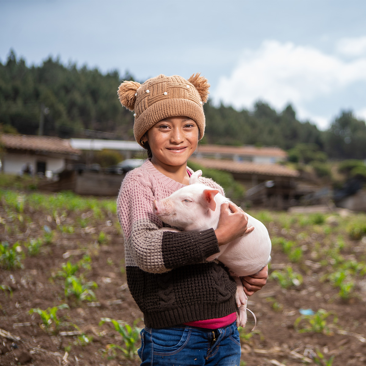 A young girl standing in a farmer’s field smiles as she holds a sleeping piglet in her arms.
