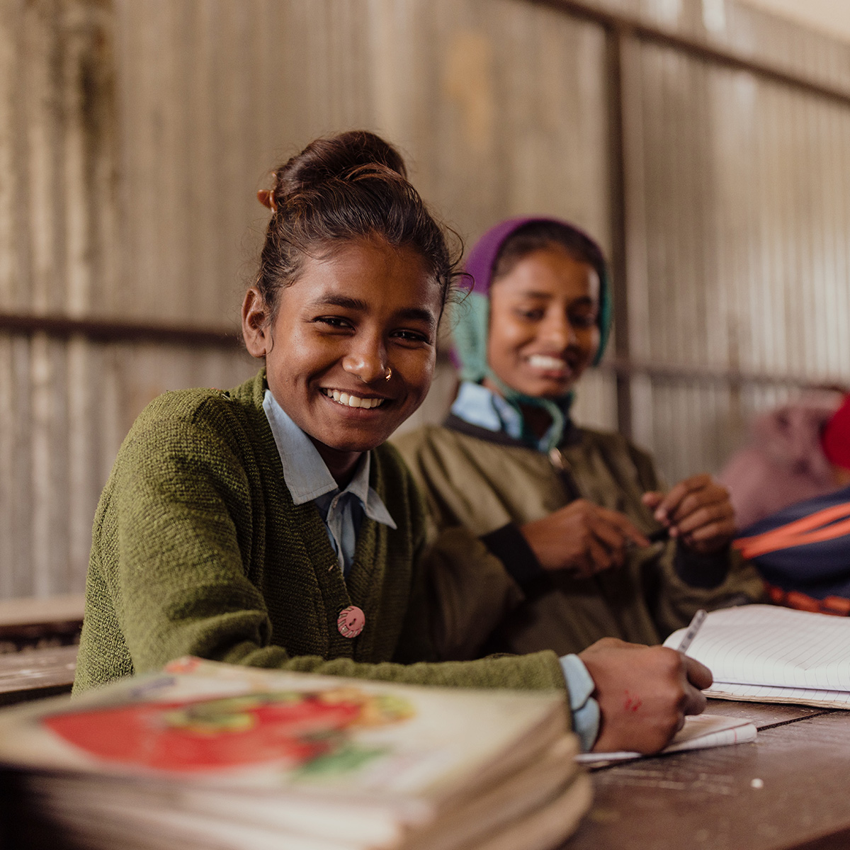 Two smiling girls sit at wooden desks in a classroom, writing in their notebooks.