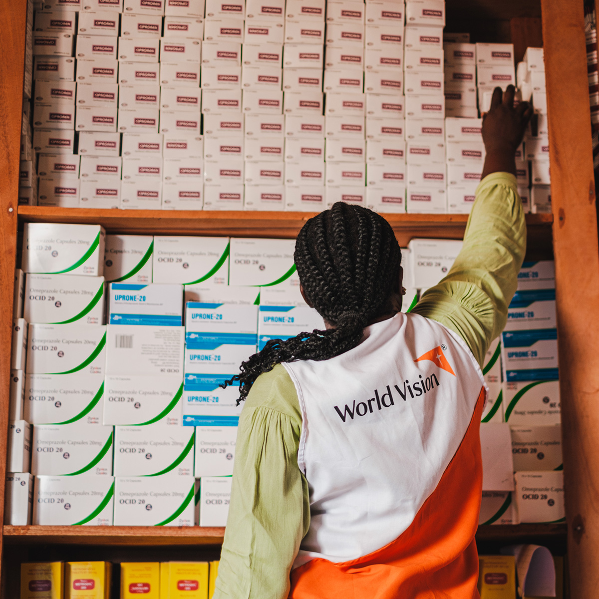 A volunteer stands inside a stocked pharmacy, reaching up  to grab medication with her back towards the camera. 
