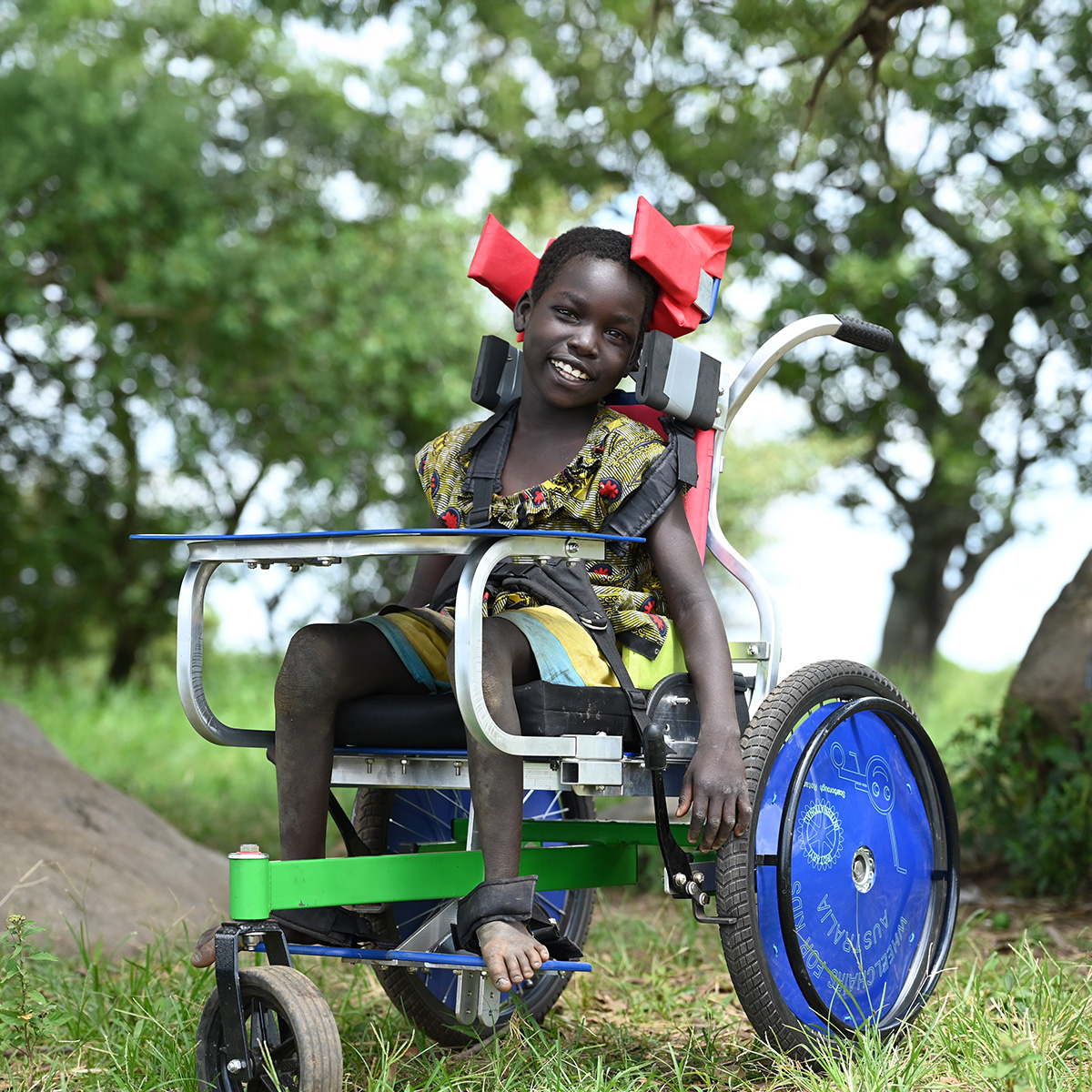 A young girl in a rural setting sits in a red, green and blue wheelchair, smiling towards the camera.