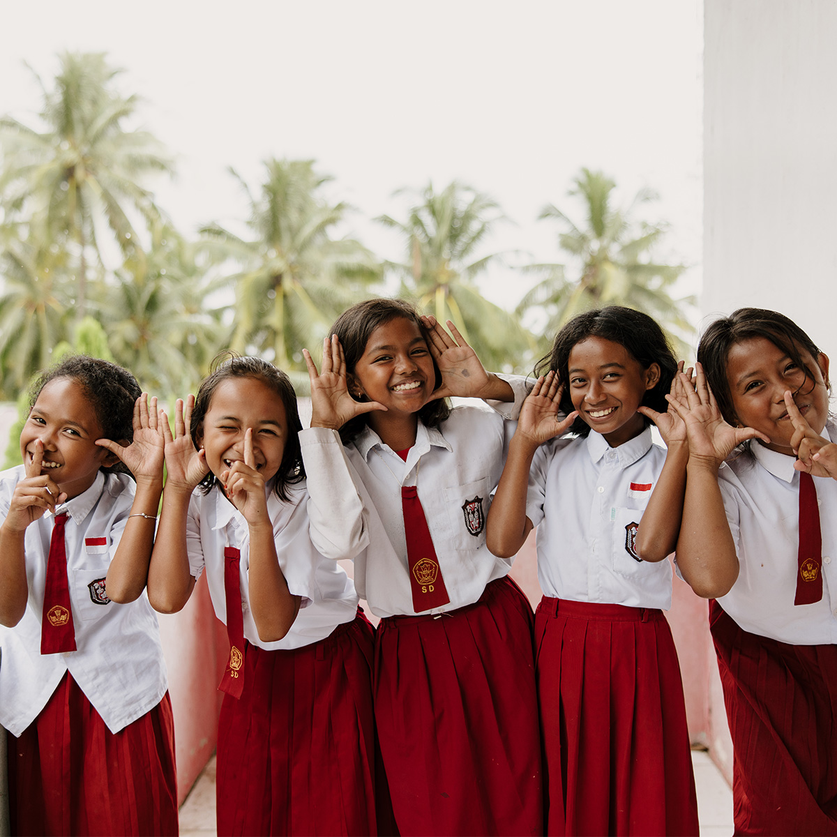 Five schoolgirls wearing burgundy skirts and ties with white blouse, smile while making playful hand gestures towards the camera.
