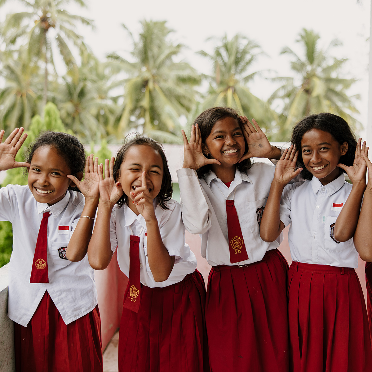 Four schoolgirls wearing burgundy skirts and ties with white blouse, smile while making playful hand gestures towards the camera.