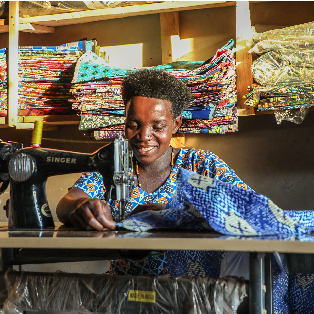 A woman is sewing patterned fabric on a black sewing machine with colorful folded textiles stacked on shelves.