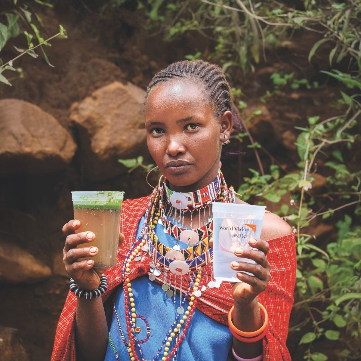 A woman wearing African clothing holds a cup of dirty water in her left hand and a cup of clean water in the other.