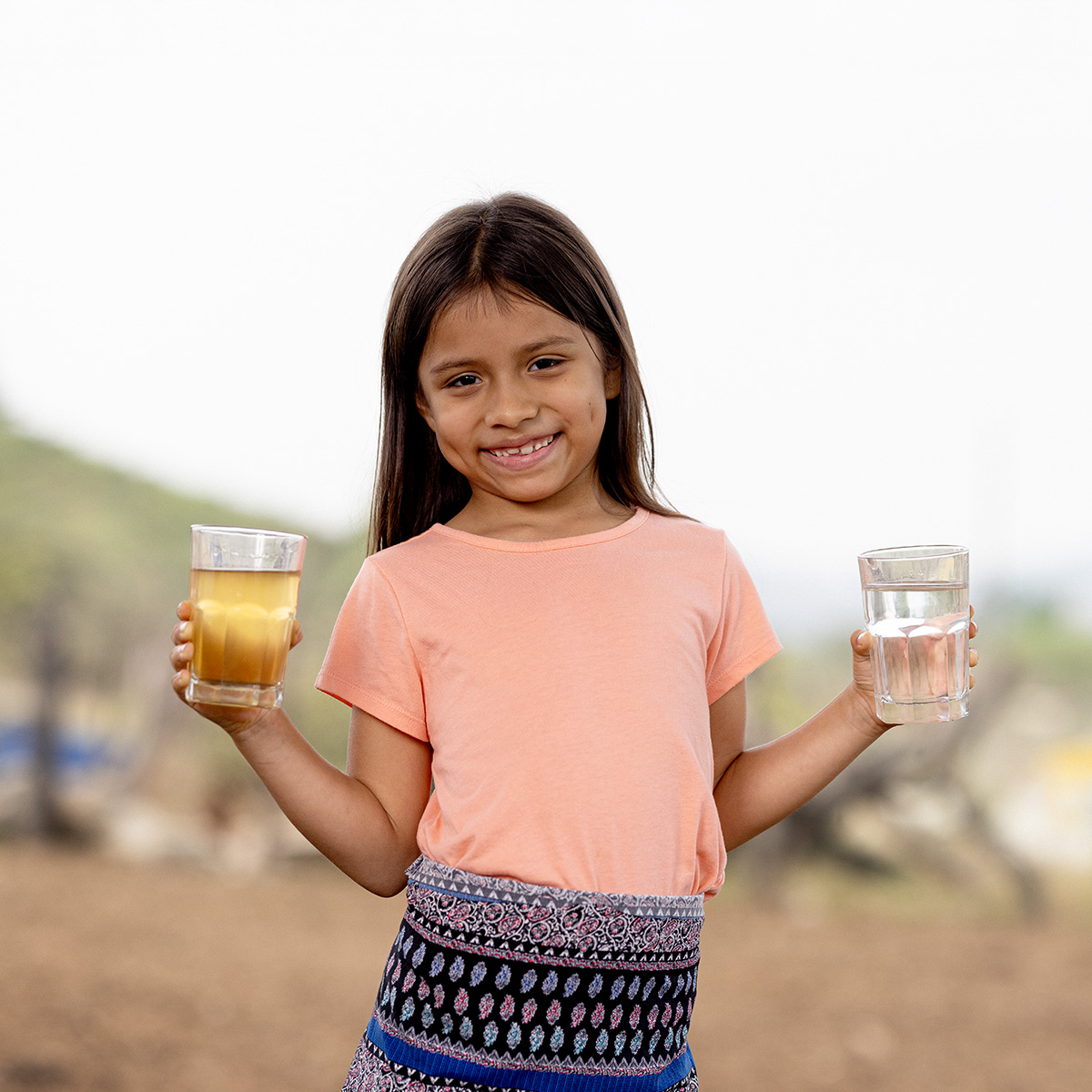 A young girl in a pink top and colourful skirt holds up two glasses of water in her hands, one clean and one dirty.