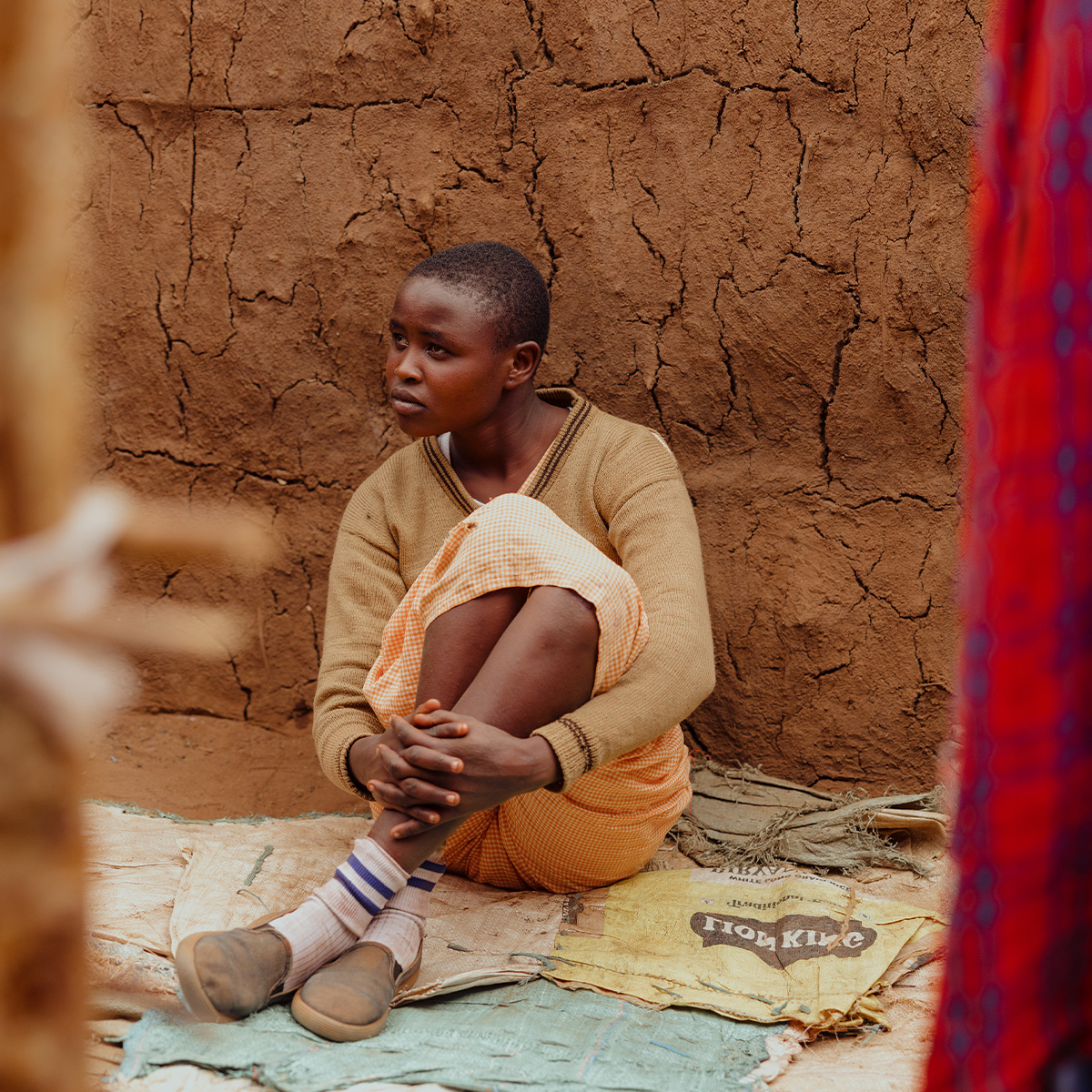 A girl sits in distress sits on old rice bags on a dirt floor, hugging her knees to her chest.