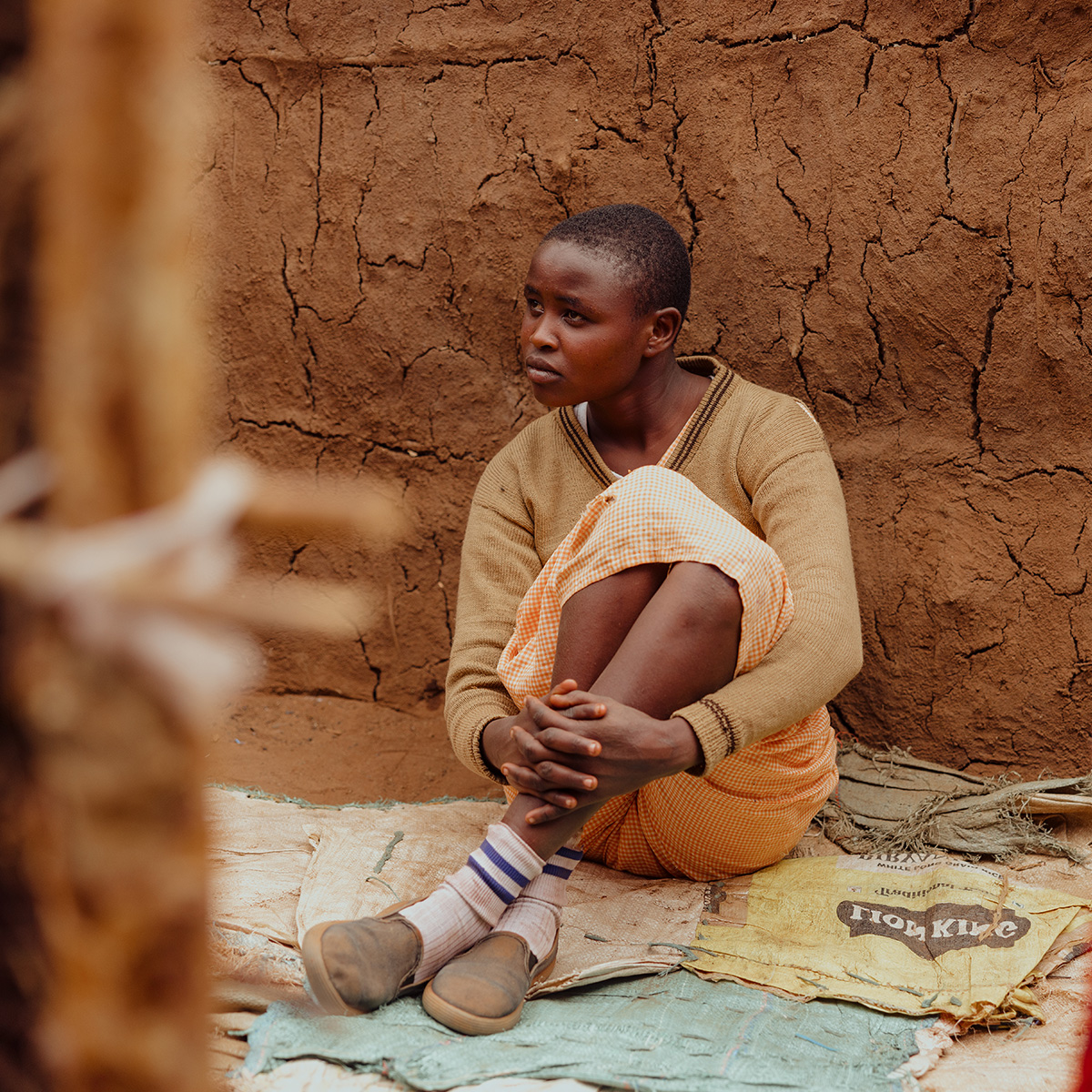 A girl sits in distress sits on old rice bags on a dirt floor, hugging her knees to her chest.