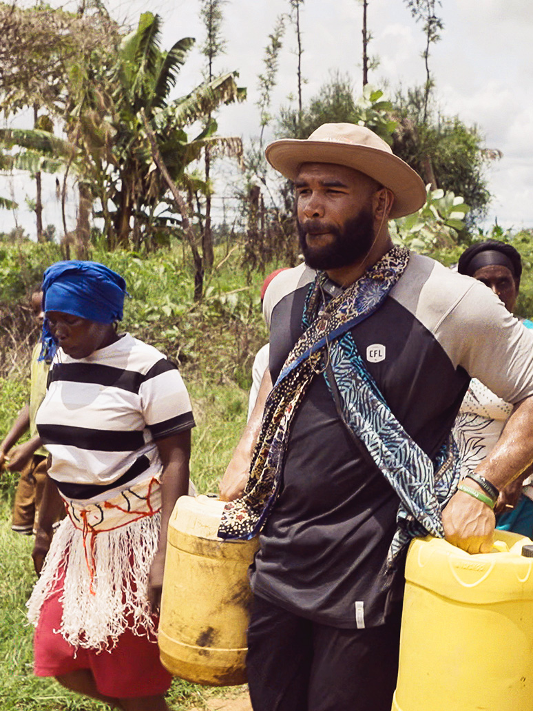 A CFL player wearing a hat, walks beside a woman as he carries two yellow buckets filled with water.