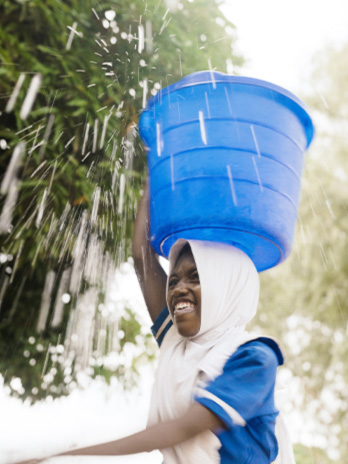 A young girl carries a blue bucket full of water on top of her head.