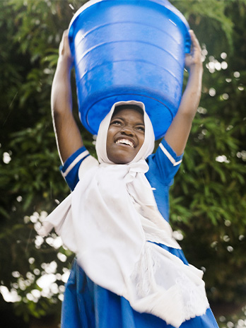 A young girl smiling while carrying a blue bucket filled with water, on top of her head.