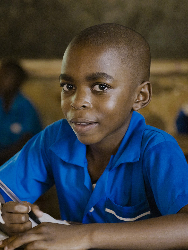 A young boy in a blue school uniform holds a pen while sitting at a desk.