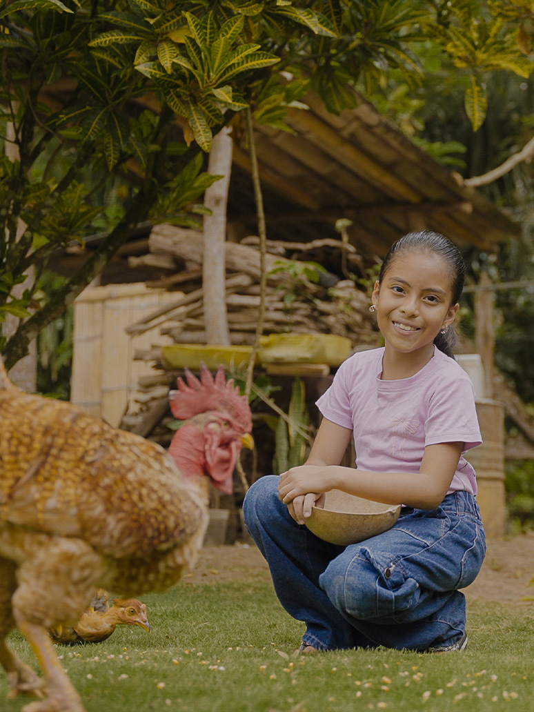 Young girl kneels behind her chicken with a bowl of feed.