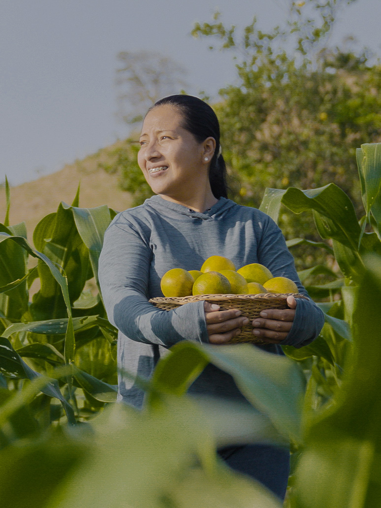 Woman smiles in a farm field, while holding a basket of oranges.