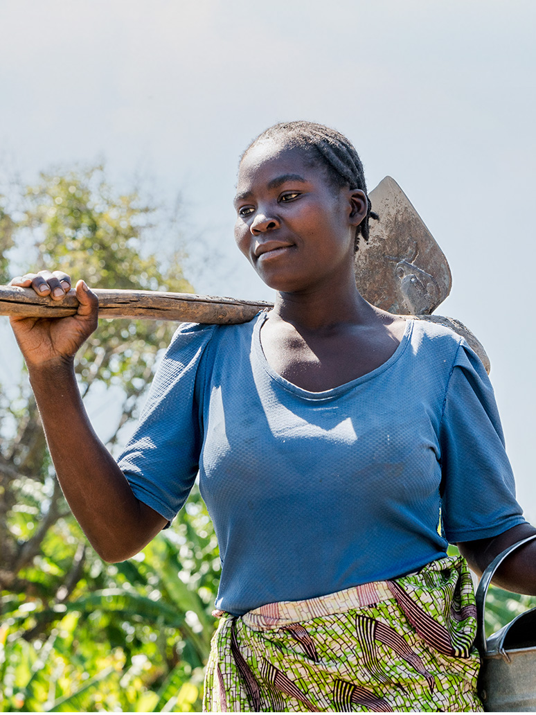 Woman smiles in a farm field while carrying a hoe on her back.